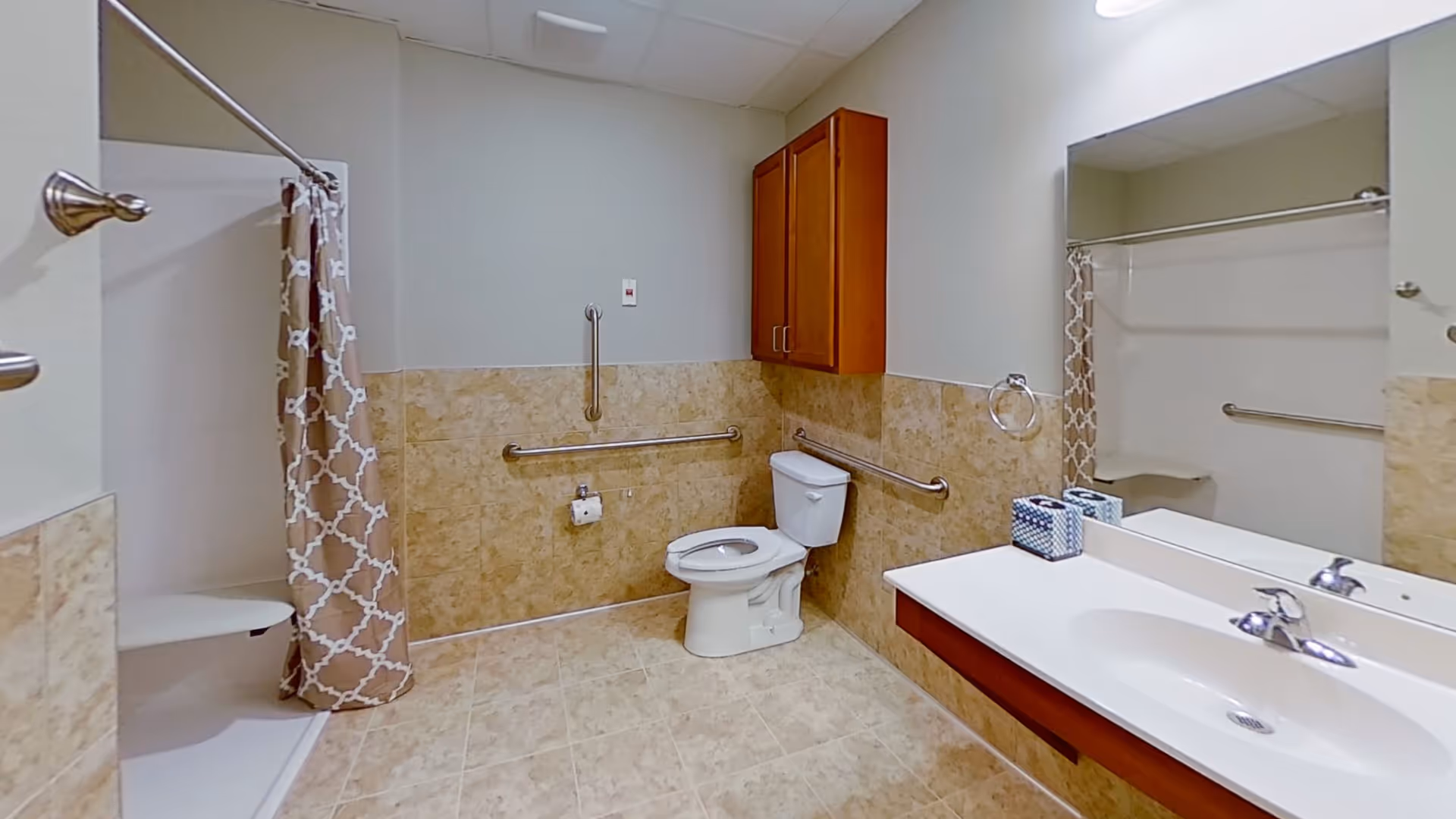 A spacious bathroom with beige tiled floor and walls halfway up. It features a white toilet with grab bars on the walls around it, a shower area with a beige and white patterned curtain, a large mirror above a white sink with a wooden cabinet below, and a wooden wall cabinet above the toilet.