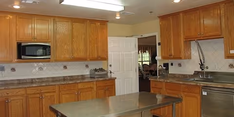 A kitchen with wooden cabinets, a microwave built into the cabinetry, a stainless steel sink, and a dishwasher. There is a metal table in the center of the room and a door leading to another room in the background.