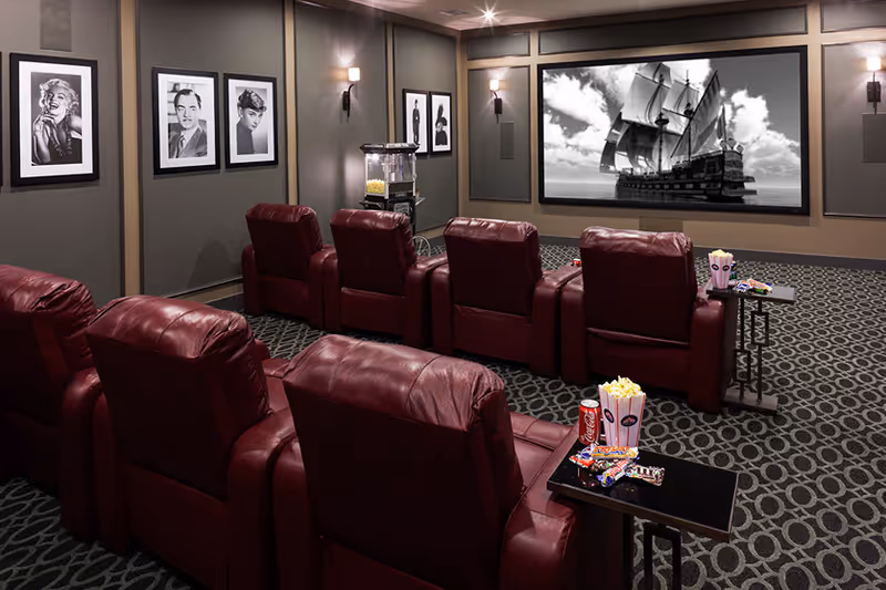 A cozy movie theater room with two rows of red leather recliner chairs facing a large screen showing a black and white image of a sailing ship. The room has patterned carpet, framed black and white celebrity photos on the walls, a popcorn machine, and small tables with popcorn, candy, and a soda can.