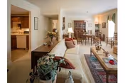 Interior view of a living room and dining area in a retirement community. The living room features a beige sofa with red cushions, a wooden coffee table on a patterned rug, and various decorative items including plants and lamps. The dining area has a wooden dining table with chairs and a china cabinet in the background. The kitchen is partially visible through an open doorway on the left.