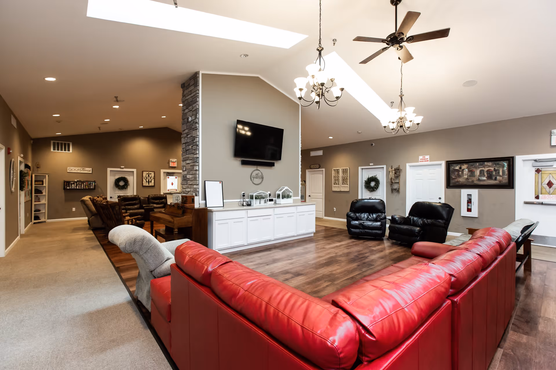 Spacious senior living facility common area with a large red sectional sofa, two black recliners, a piano, and a wall-mounted TV. The room features wood flooring, beige walls, ceiling fans, chandeliers, and decorative wreaths on the doors.