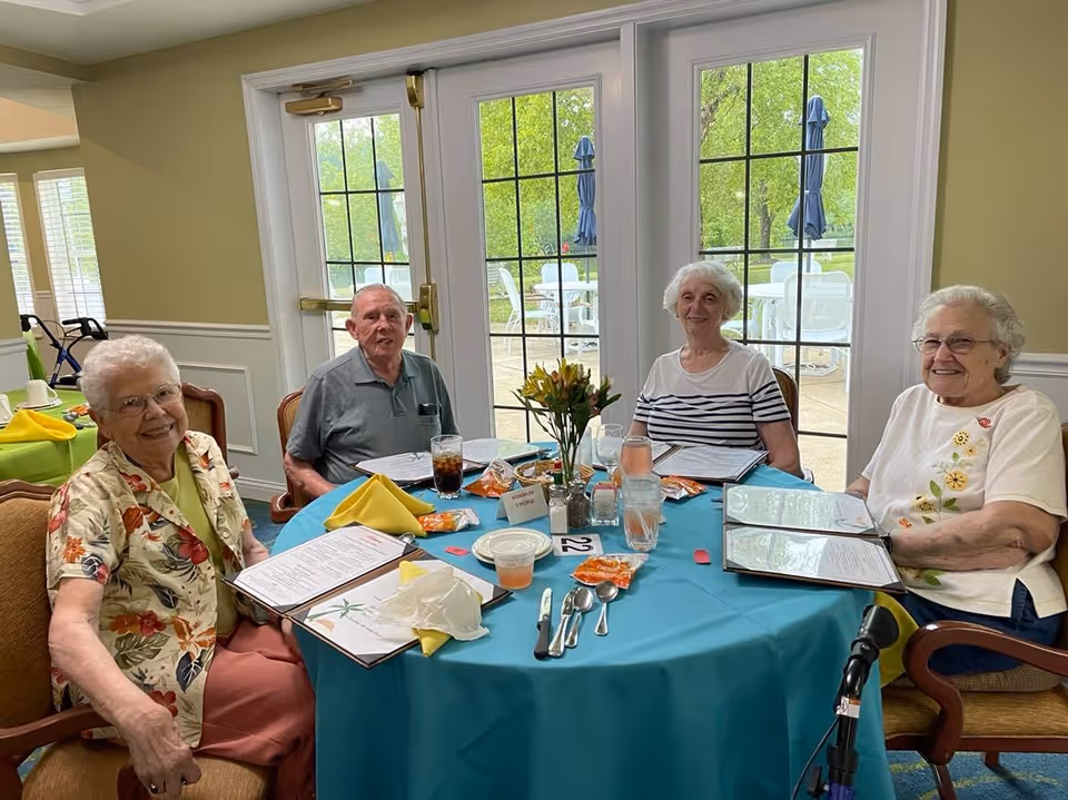 Four elderly individuals sitting around a round table covered with a teal tablecloth in a dining area. They have menus in front of them and are smiling. The table is set with utensils, glasses, napkins, and condiment packets. Behind them are large glass doors showing an outdoor patio with tables and umbrellas.