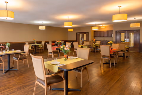 A spacious dining room with multiple tables and chairs arranged neatly. Each table has a small flower arrangement and place settings. The room features warm lighting from ceiling lamps and has wooden flooring and beige walls with dark wood paneling along the lower half.