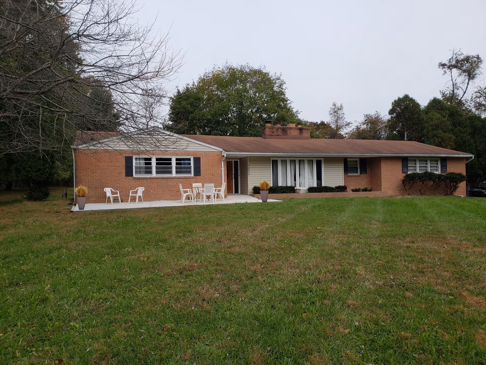 Single-story brick and siding building with a brown roof, surrounded by green grass and trees. There is a concrete patio area with white chairs and a table, along with potted plants on either side of the patio.