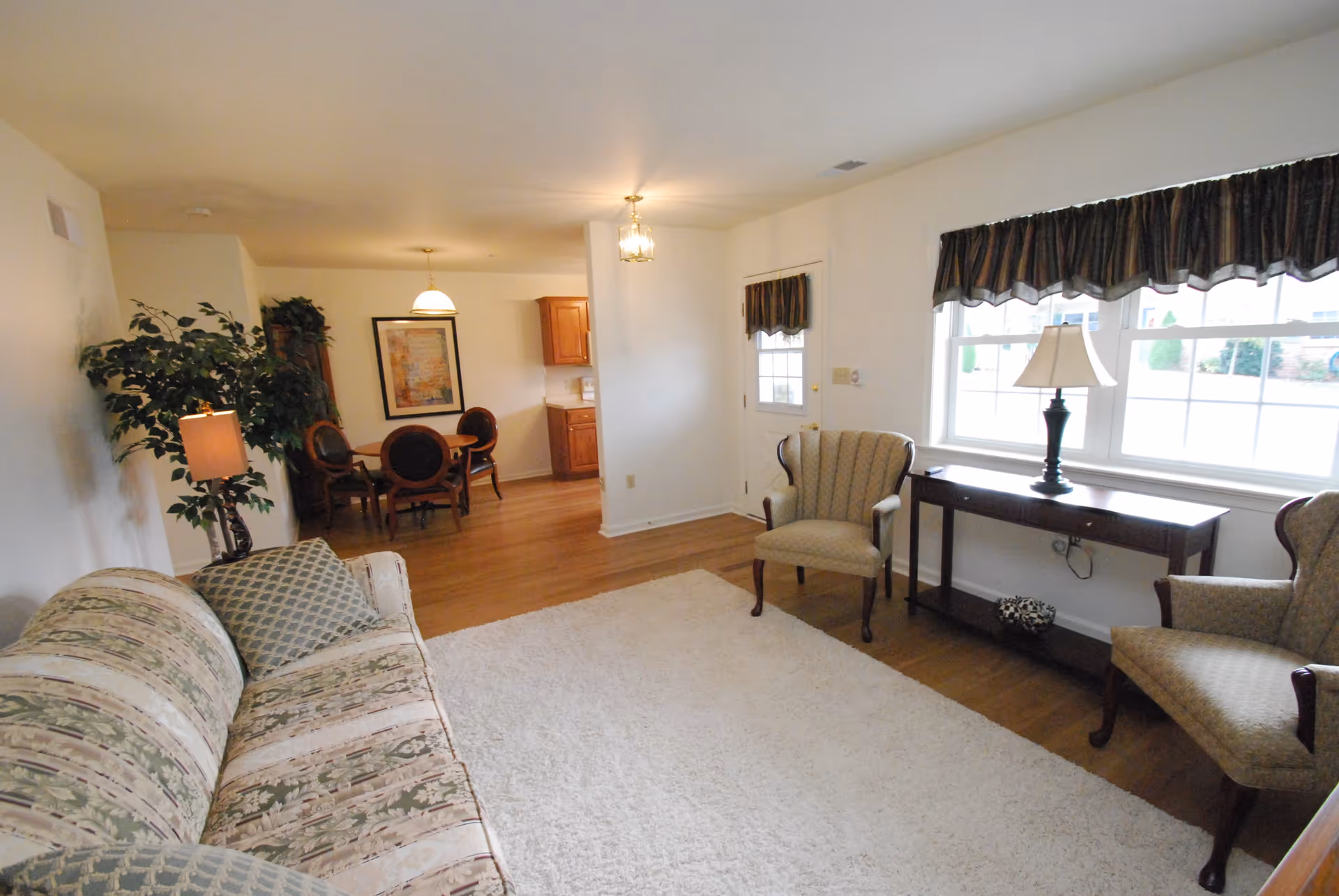 A cozy living room with a patterned sofa, two armchairs, a wooden console table with a lamp, and a large window with dark valances. The room has hardwood floors and a light-colored area rug. In the background, there is a small dining area with a round table and four chairs, a framed picture on the wall, and a kitchen area with wooden cabinets.
