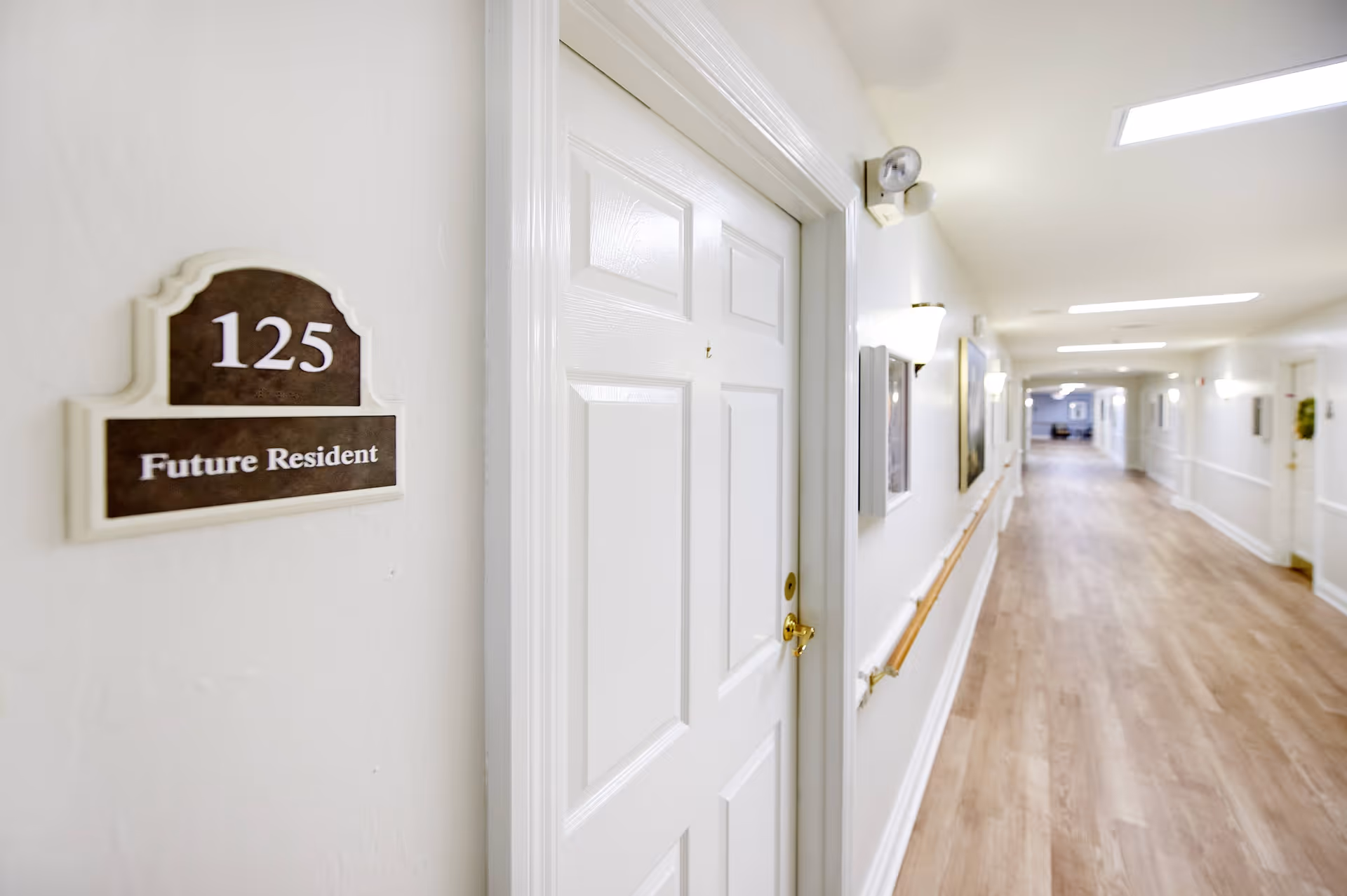 A bright hallway in a senior living facility with light wood flooring and white walls. A white door labeled '125 Future Resident' is visible on the left side. The hallway has handrails along the walls and several light fixtures, with artwork hanging on the walls in the distance.