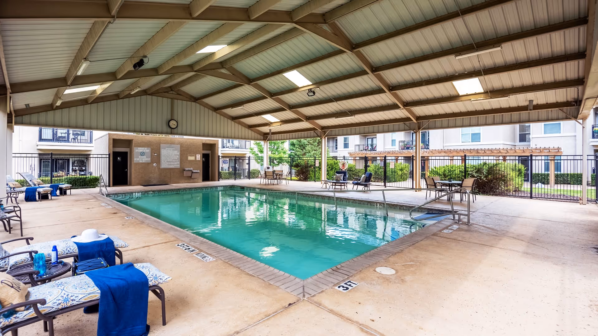 Covered outdoor swimming pool area with lounge chairs and tables around it. The pool is surrounded by a concrete deck and a metal fence. In the background, there are apartment-style buildings and greenery.