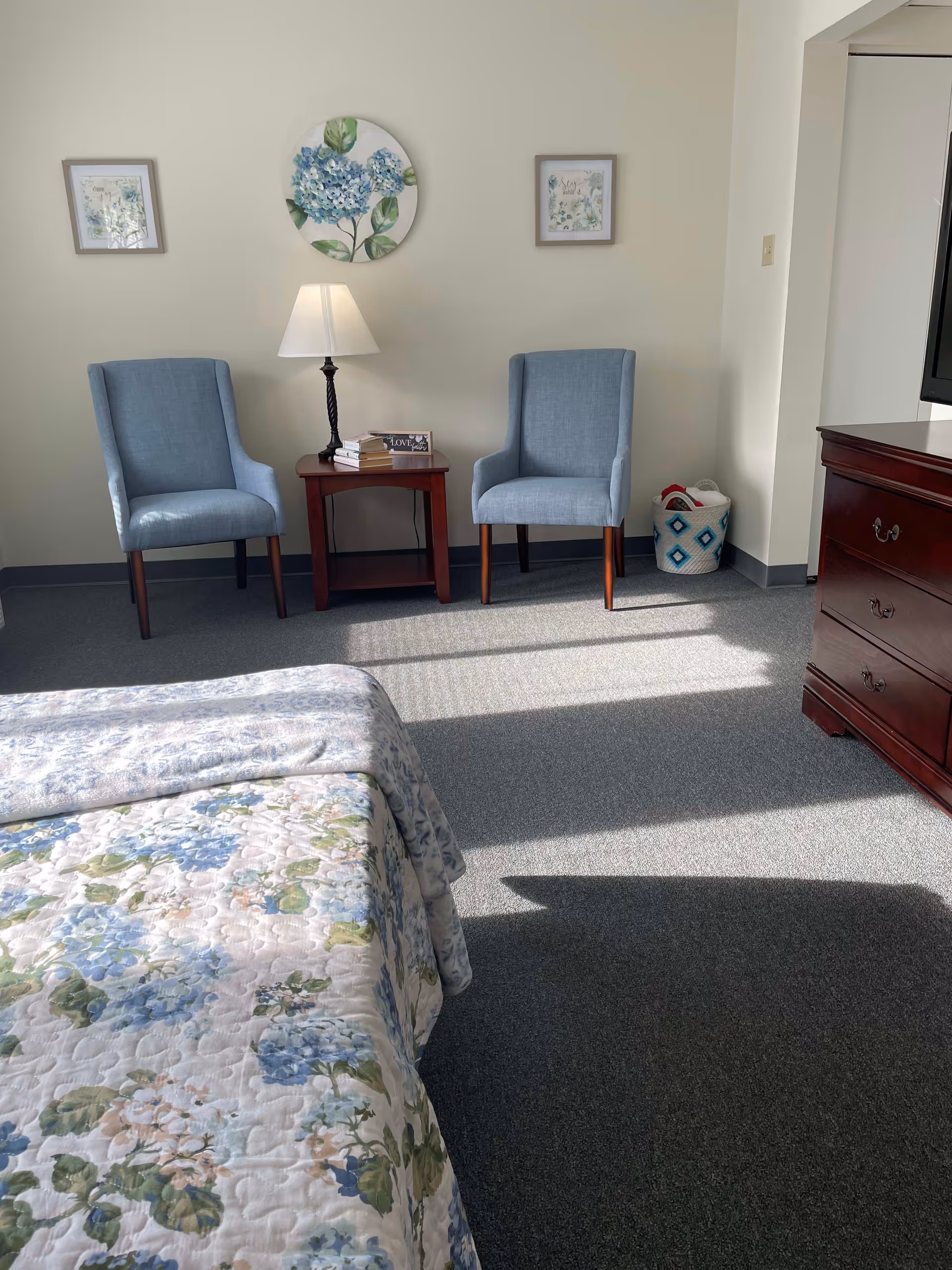 A bedroom with two twin beds covered in floral bedspreads, two blue upholstered chairs with wooden legs, a small wooden side table with a lamp and books, floral artwork on the wall, a basket in the corner, and a wooden dresser with a TV on top.