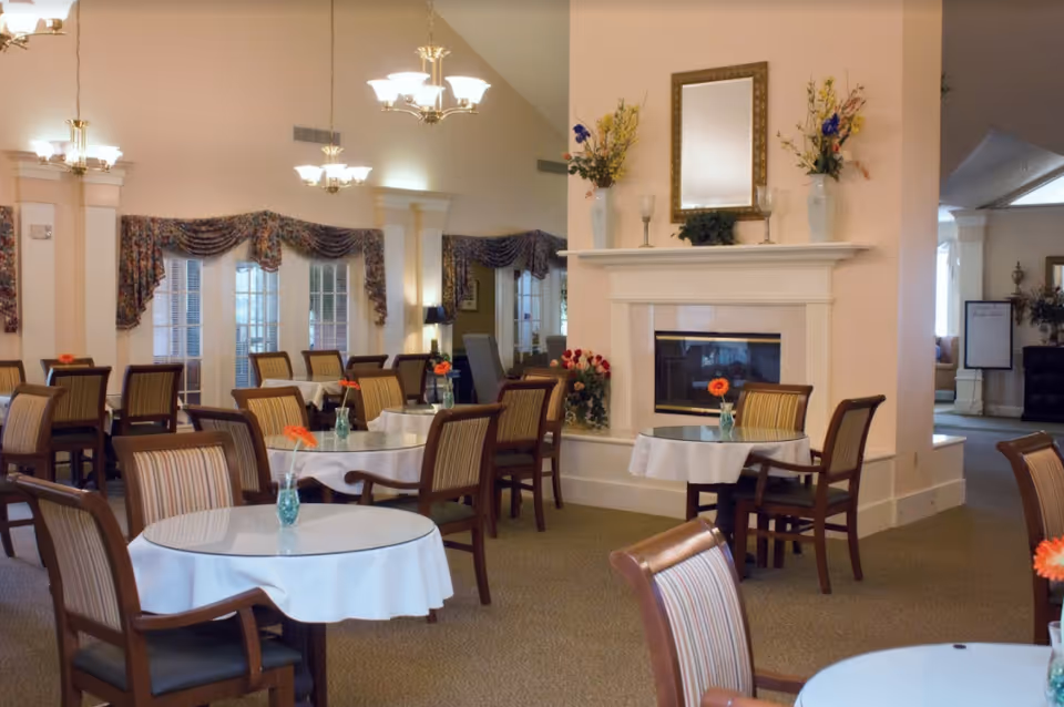 A dining room with several round tables covered with white tablecloths, each table having a small vase with an orange flower. The room features wooden chairs with striped cushions, a fireplace with a mirror and floral arrangements on the mantel, and large windows with decorative curtains. The lighting is provided by elegant chandeliers hanging from the ceiling.
