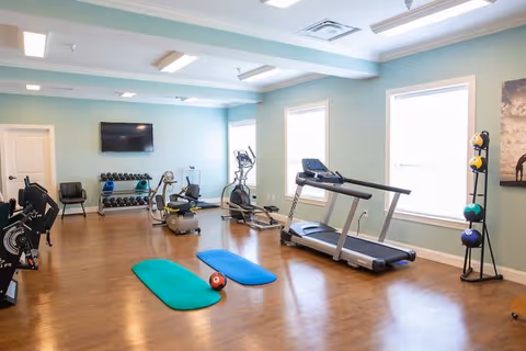 A well-lit fitness room with wooden flooring featuring exercise equipment including a treadmill, stationary bikes, elliptical machine, two exercise mats, a medicine ball, and a rack of dumbbells. The room has light blue walls, three large windows, and a wall-mounted flat screen TV.