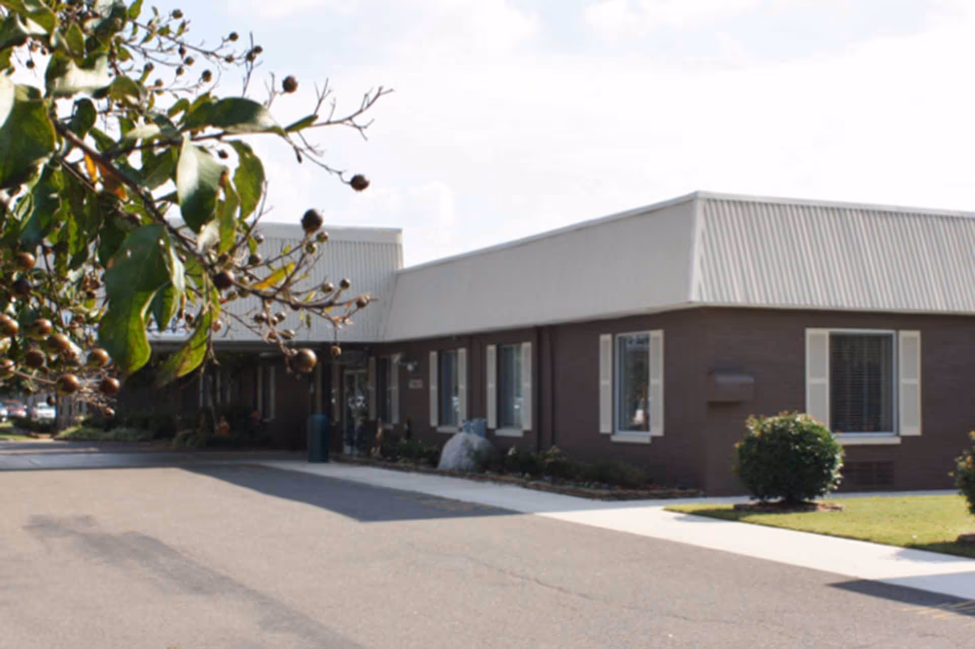 Exterior view of a single-story building with a light-colored roof and brown walls, surrounded by a paved driveway and some greenery including bushes and a tree with leaves and small round fruits in the foreground.