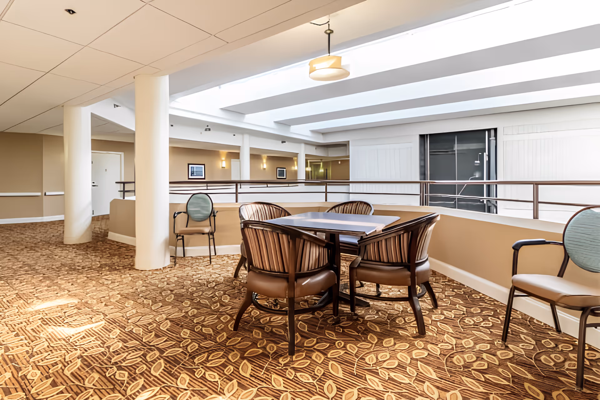 A bright indoor seating area in Elison Independent Living of Niles featuring a square table surrounded by four cushioned chairs with striped upholstery. The space has a patterned carpet with leaf designs, beige walls, white ceiling with recessed lighting, and large skylights allowing natural light to fill the area. Additional chairs are placed along the wall, and the area overlooks a lower floor through a railing.