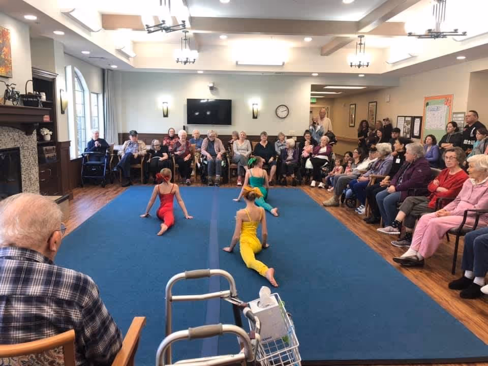 A group of elderly residents seated in chairs around a large blue carpet in a common room, watching three young girls in colorful dance costumes performing on the carpet. The room has wooden flooring, a fireplace on the left, a wall-mounted TV, and several light fixtures on the ceiling.