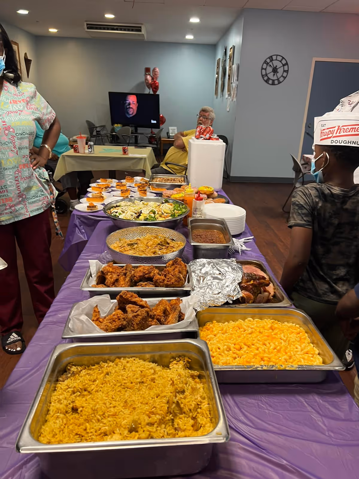 A buffet table covered with a purple tablecloth displaying trays of food including yellow rice, macaroni and cheese, fried chicken, salad, baked beans, and other dishes. In the background, a person wearing a Krispy Kreme doughnuts hat and a face mask stands near the table. Another person in scrubs stands to the left. A TV screen and a seated elderly person are visible in the room.