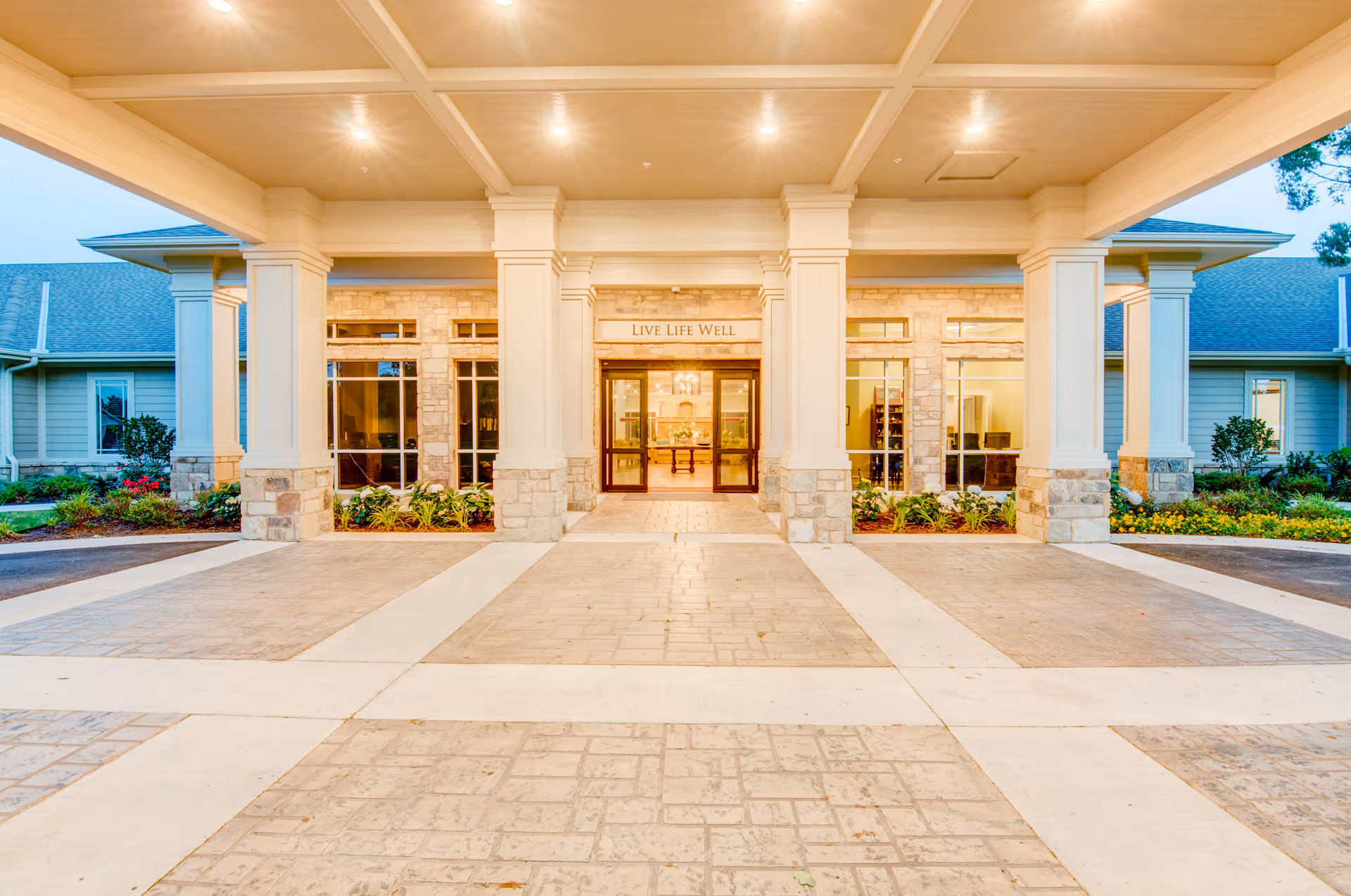 Entrance of Village on the Park Bentonville facility with a covered driveway supported by large white columns, stone accents on the building, well-lit ceiling, and landscaping with plants and flowers on either side.