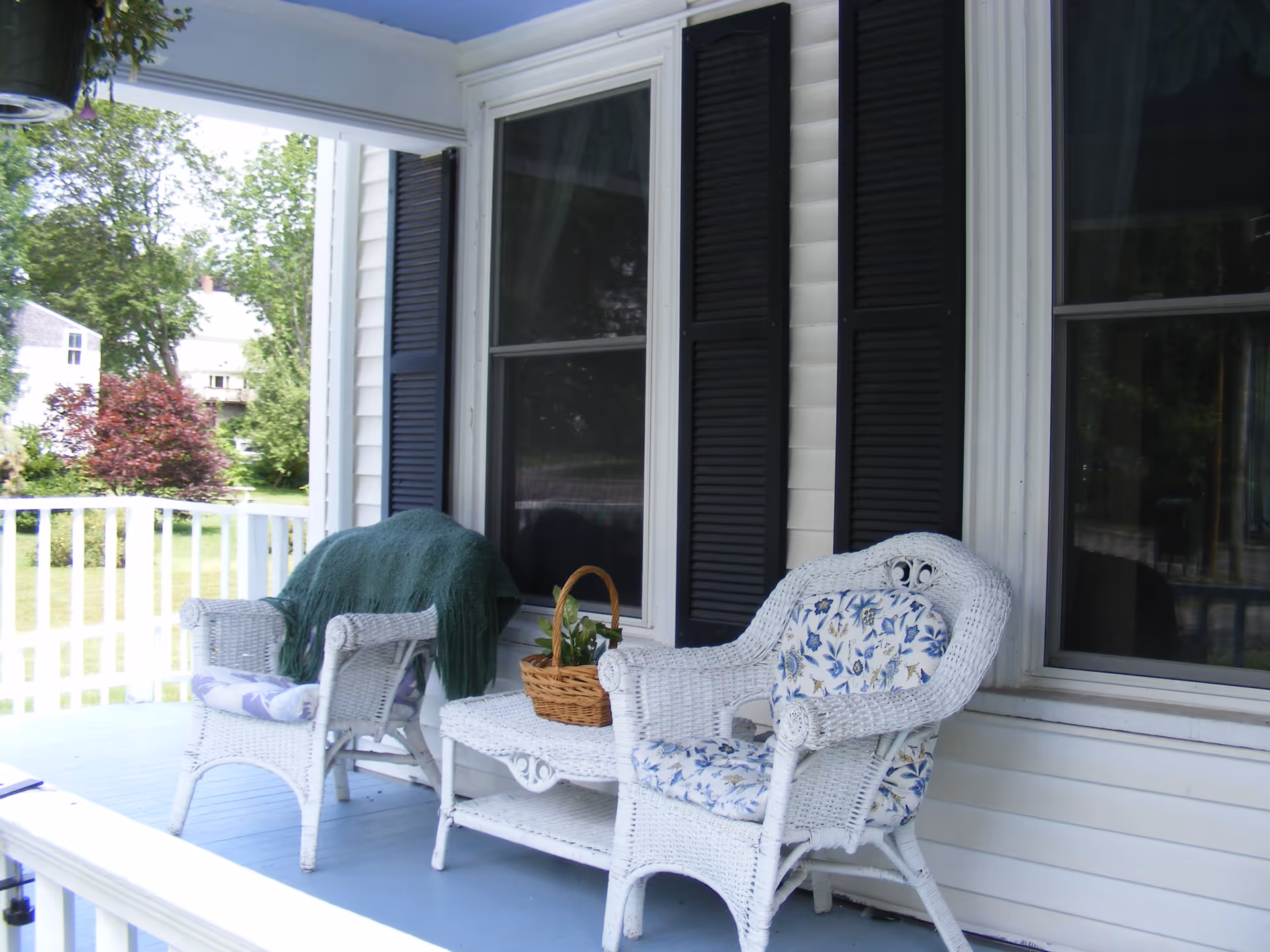 A porch area with two white wicker chairs with floral cushions and a small white wicker table between them. One chair has a green blanket draped over it and there is a basket with plants on the table. The porch has white railings and is attached to a house with white siding and black shutters on the windows. Trees and a lawn are visible in the background.