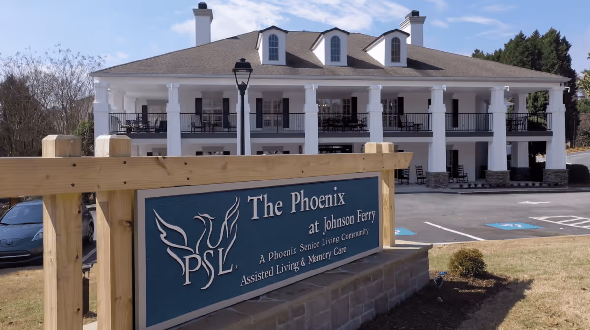 Exterior view of The Phoenix at Johnson Ferry senior living community building with a large white two-story structure featuring columns and balconies. In the foreground, there is a wooden sign with the community's name and description, along with a parking lot and some landscaping.