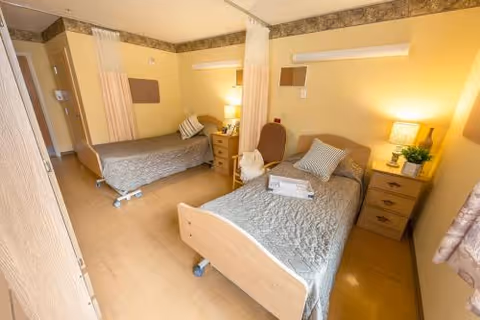 A well-lit room with two single hospital-style beds separated by a curtain. Each bed has a bedside table with a lamp and some personal items. The room has light-colored walls and flooring, with a chair placed between the beds.