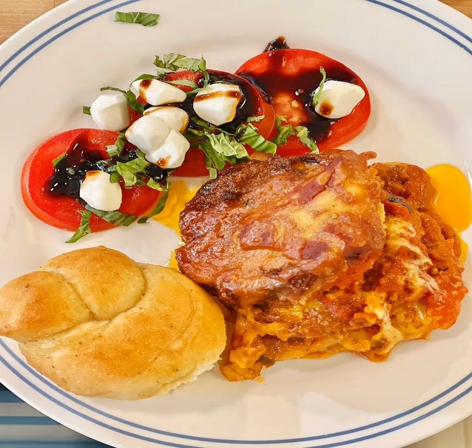 A plate with a serving of lasagna, a dinner roll, and a side salad of sliced tomatoes topped with small mozzarella balls, chopped basil, and balsamic glaze.