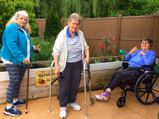 Three elderly women enjoying time outdoors in a garden area. One woman is standing with a cane, another is using a walker, and the third is sitting in a wheelchair giving a thumbs-up. Behind them is a raised garden bed with plants and a wooden fence.