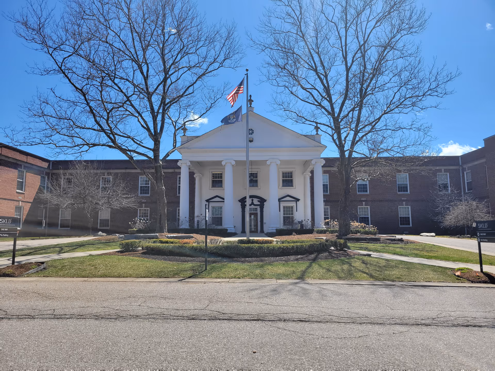 Brick senior living building with a white-columned entrance, flagpole, and leafless trees in front on a sunny day.
