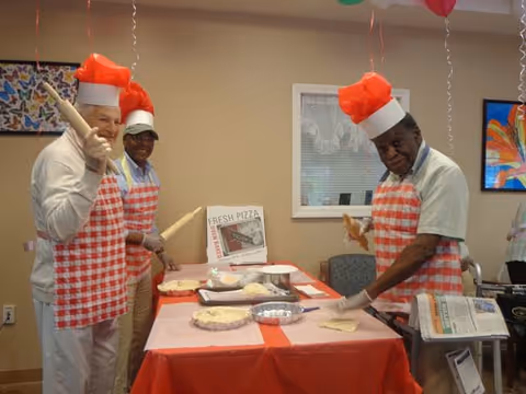 Three men wearing red and white checkered aprons and red chef hats are preparing pizzas on a table covered with a red tablecloth. They are smiling and holding rolling pins and pizza dough in a room decorated with balloons and colorful artwork on the walls.
