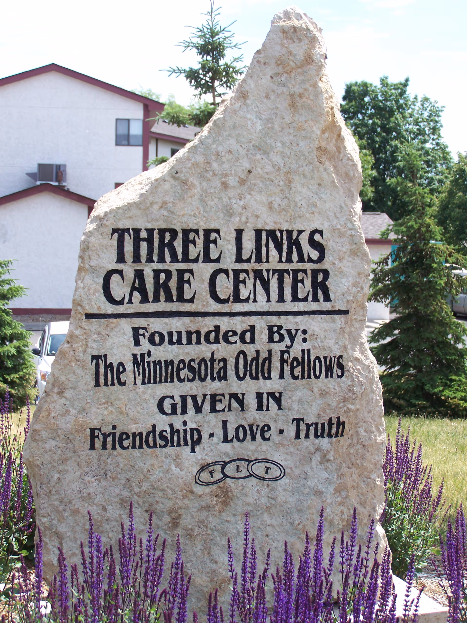 A large stone monument with the inscription 'THREE LINKS CARE CENTER Founded By: The Minnesota Odd Fellows GIVEN IN Friendship · Love · Truth' surrounded by purple flowers and greenery, with a building and trees in the background.