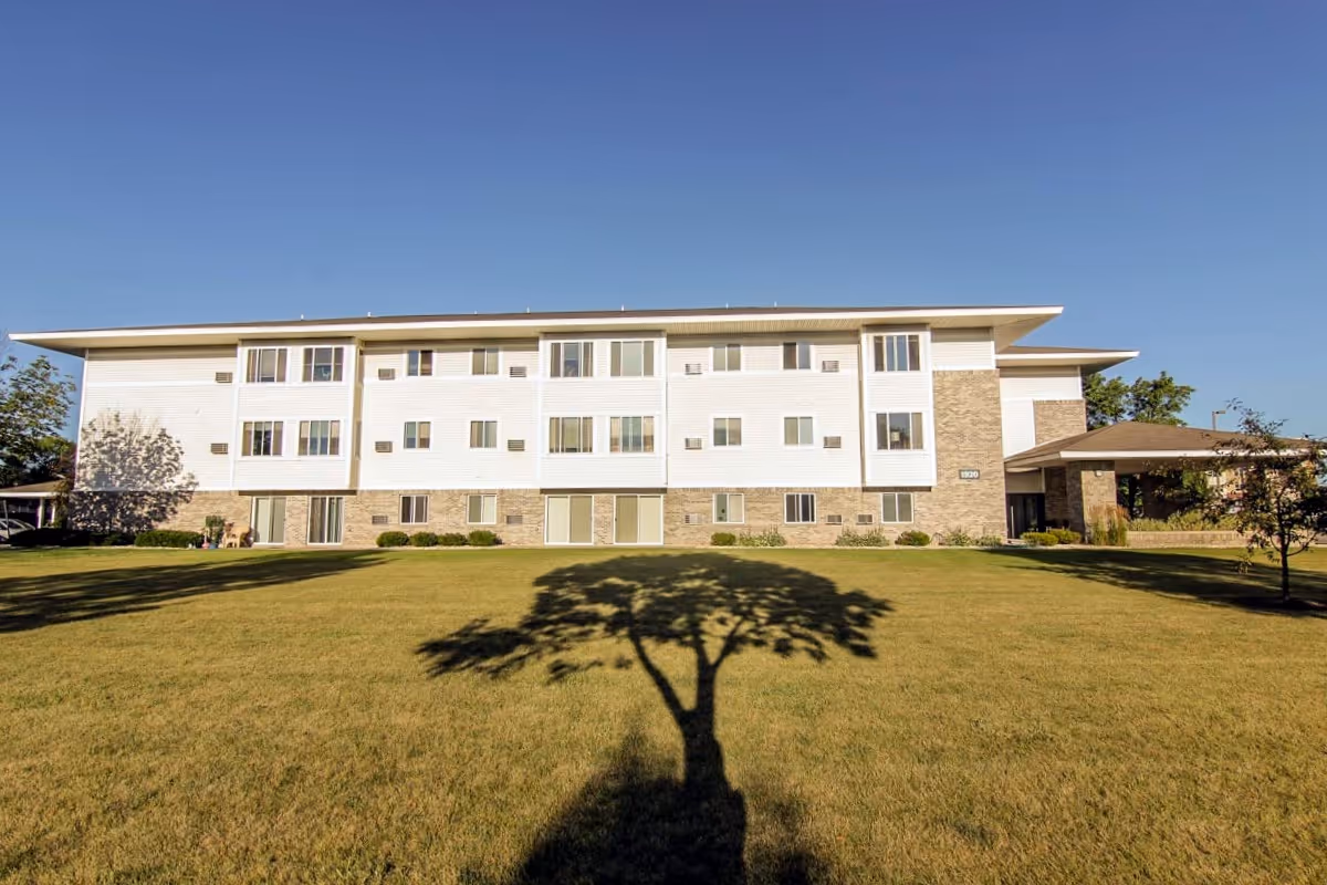 Three-story white and brick apartment building with a large grassy lawn and a tree shadow in the foreground under a clear blue sky.