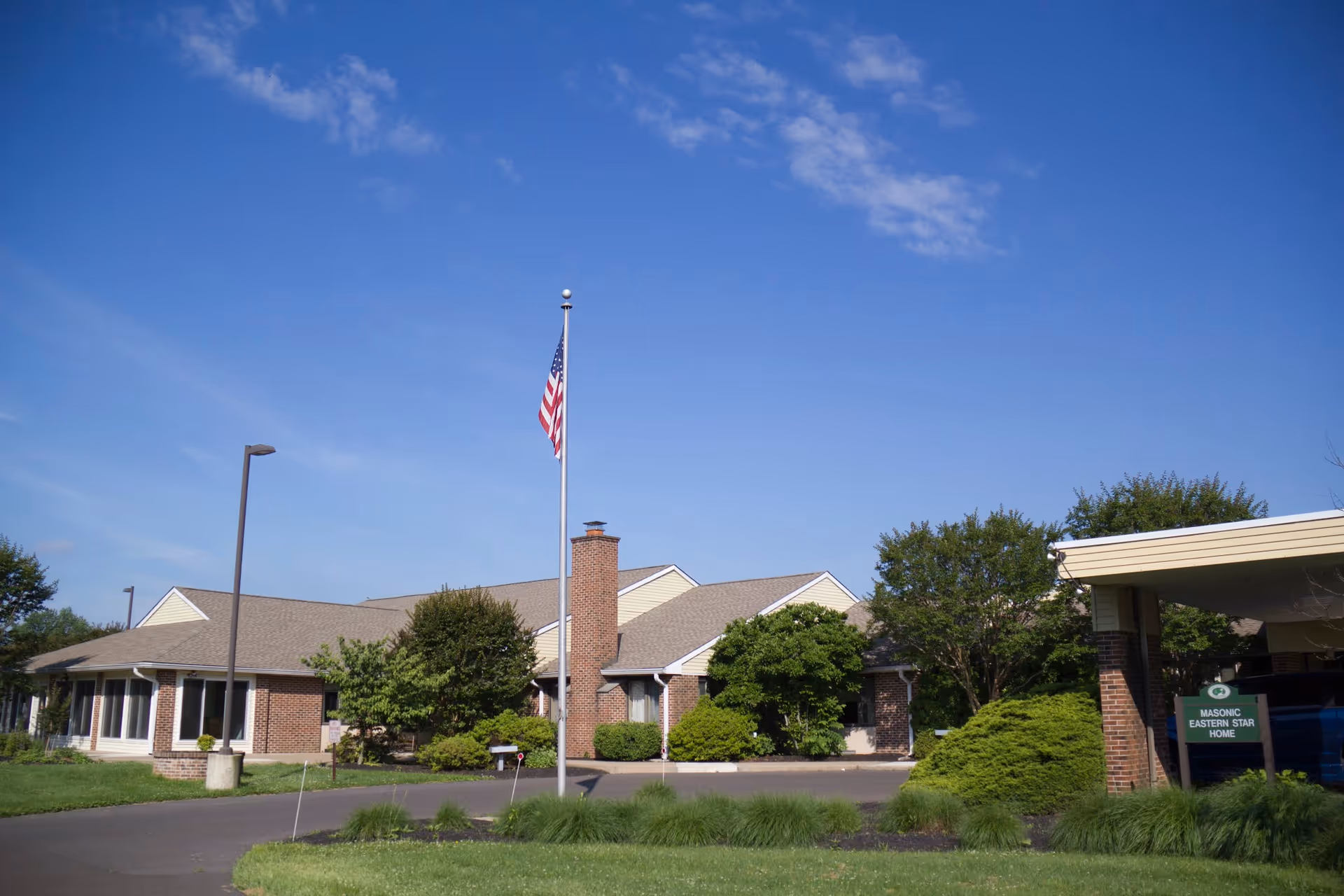 Exterior view of a single-story brick building with a chimney, surrounded by green bushes and trees under a clear blue sky. An American flag is flying on a flagpole in front of the building. A sign near the entrance reads 'Masonic Eastern Star Home.'
