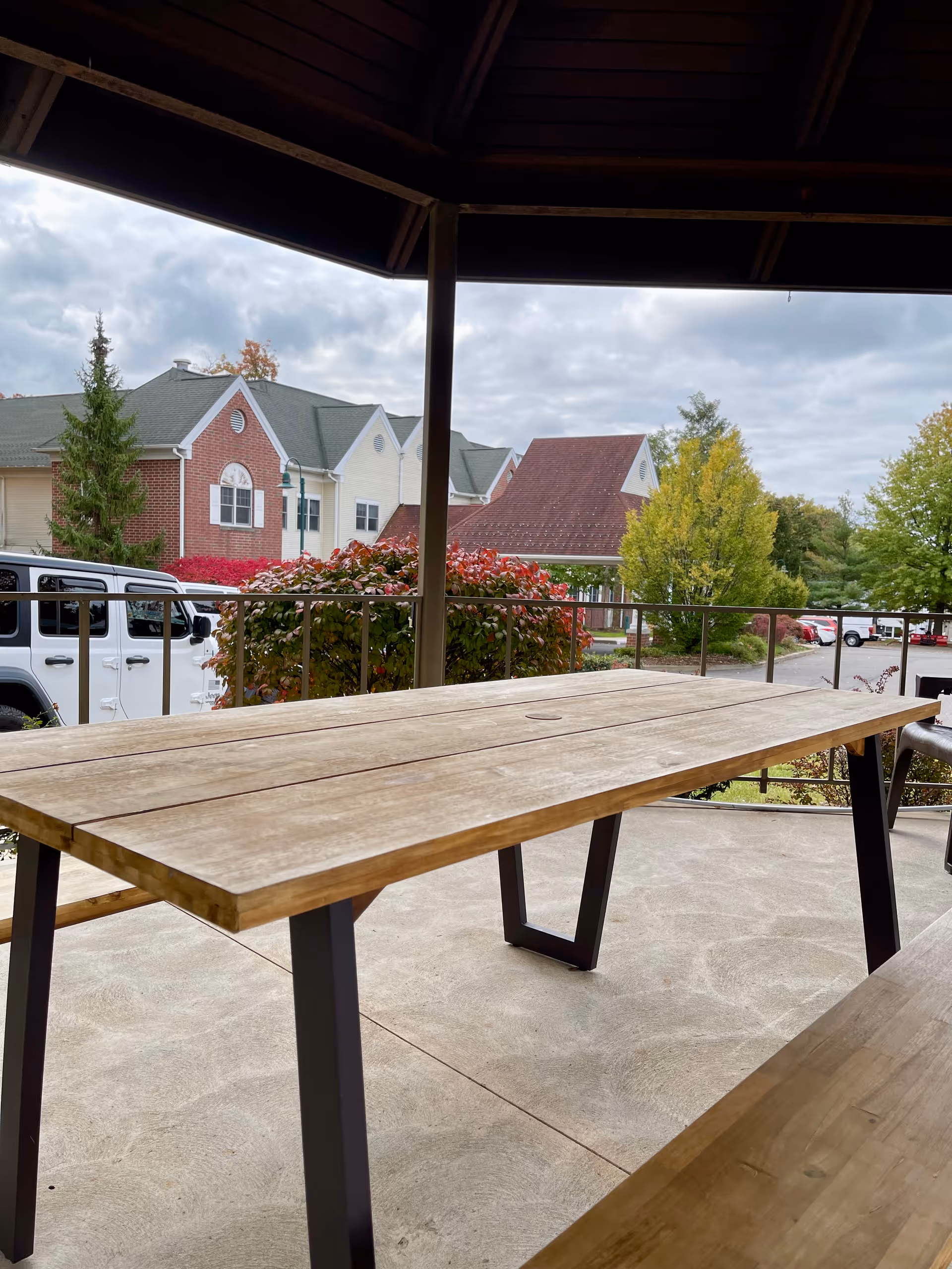Outdoor covered patio area with a wooden picnic table and benches. In the background, there are bushes, trees, parked cars, and residential-style buildings with brick and siding exteriors under a cloudy sky.