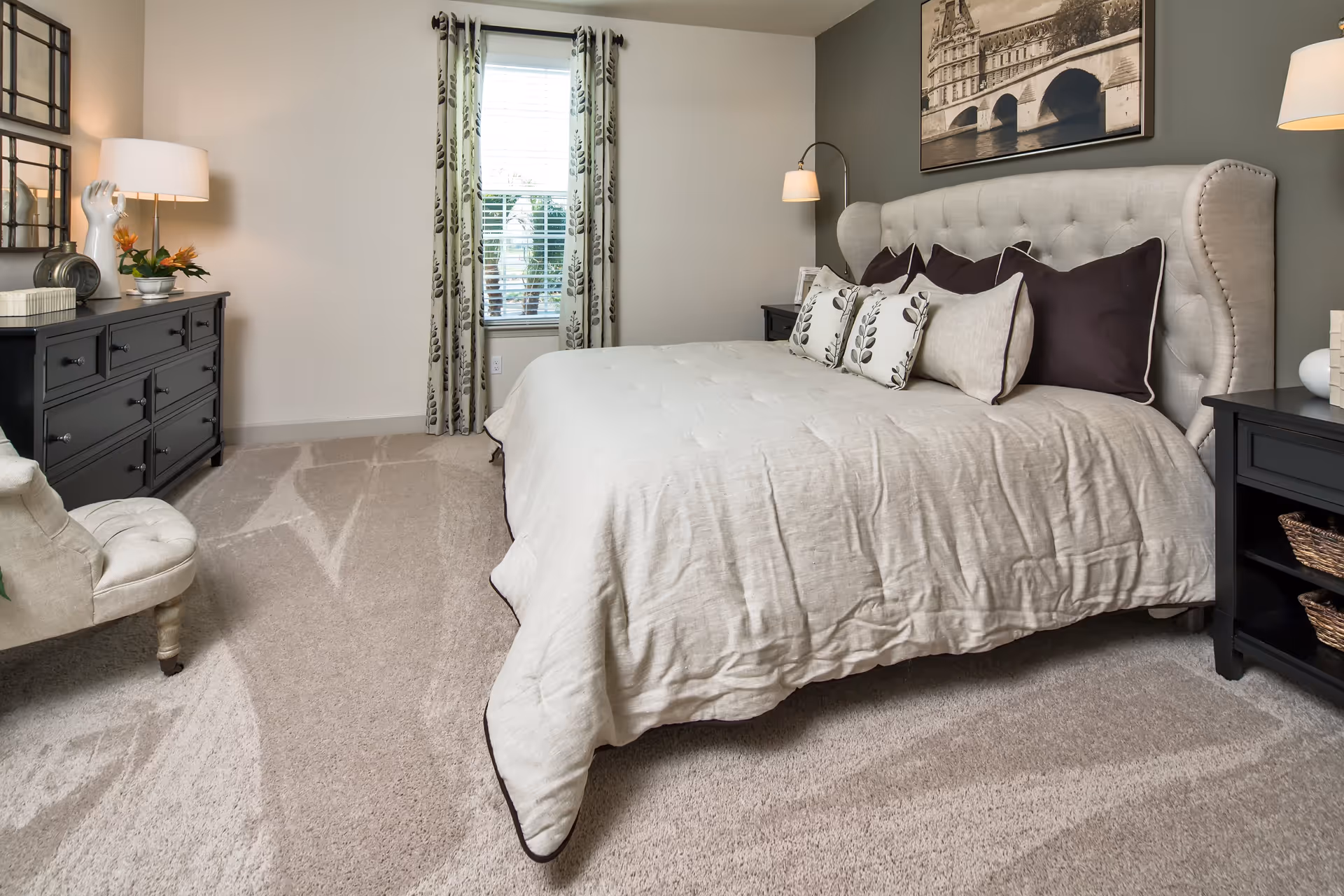 A neatly arranged bedroom with a large bed featuring a tufted headboard and multiple pillows. The room has beige carpeting, a window with patterned curtains, a black dresser with a lamp and decorative items, and a cushioned chair. A framed black and white picture hangs above the bed.