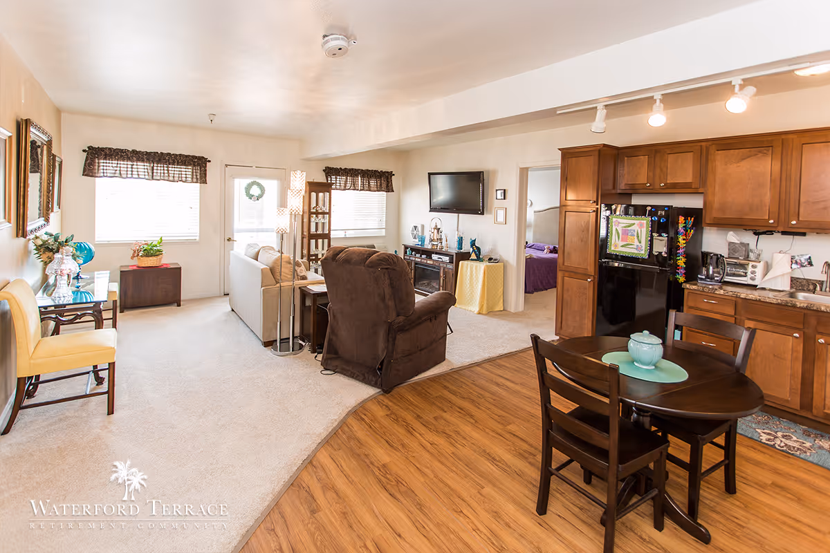 A bright and cozy retirement community apartment featuring an open living room and kitchen area. The living room has a beige sofa, a brown recliner, a TV mounted on the wall, and decorative items. The kitchen area includes wooden cabinets, a black refrigerator, a small round dining table with two chairs, and various kitchen appliances. There is a doorway leading to a bedroom with a purple bedspread. The floor transitions from carpet in the living area to wood laminate in the kitchen. Windows with dark valances let in natural light.