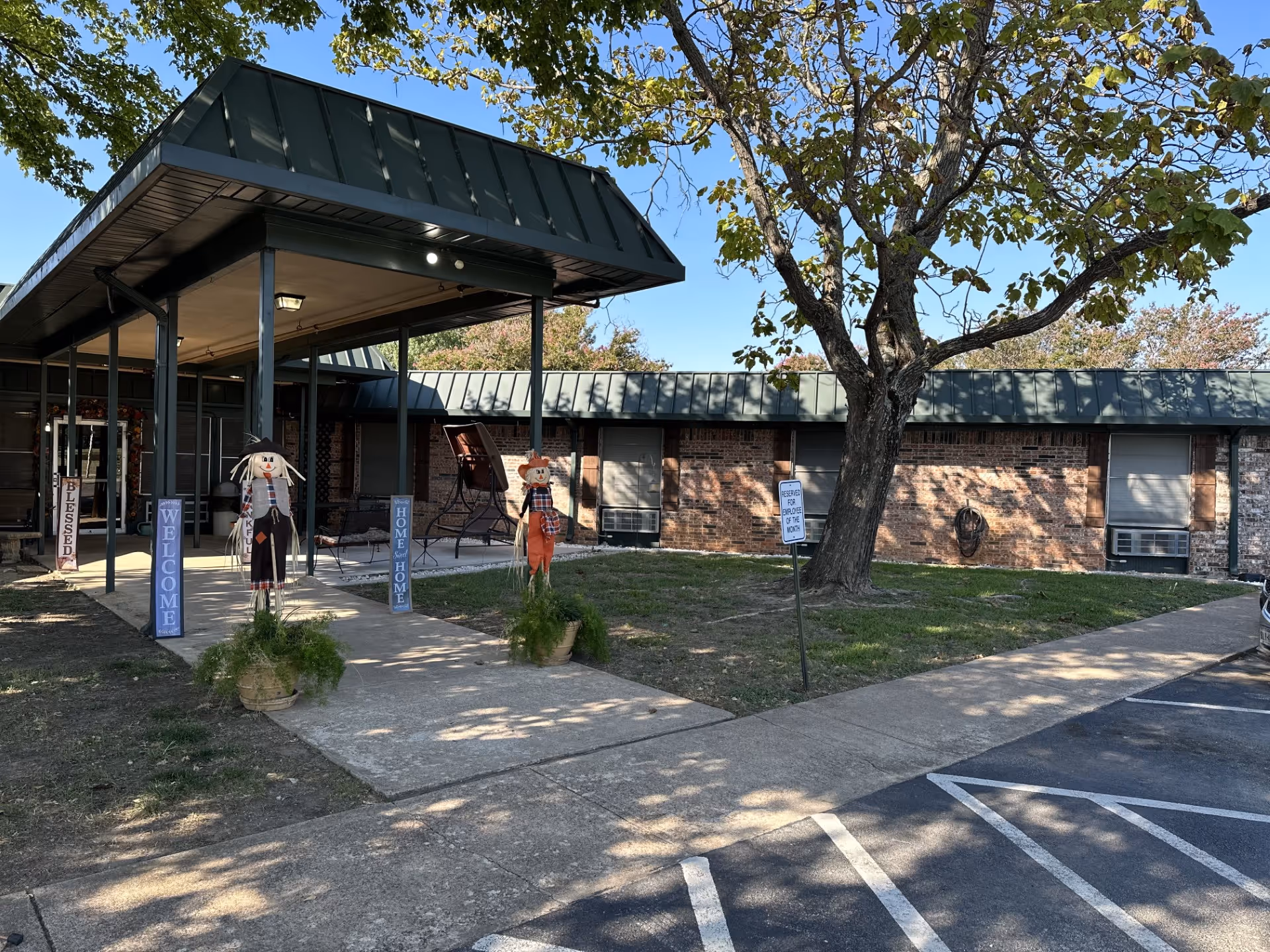 Exterior view of Pecan Creek Healthcare Center showing a covered entrance with a green metal roof, two scarecrow decorations in pots, and signs that say 'Blessed', 'Welcome', and 'Home Home'. There is a tree near the entrance and a parking area with marked spaces in front.