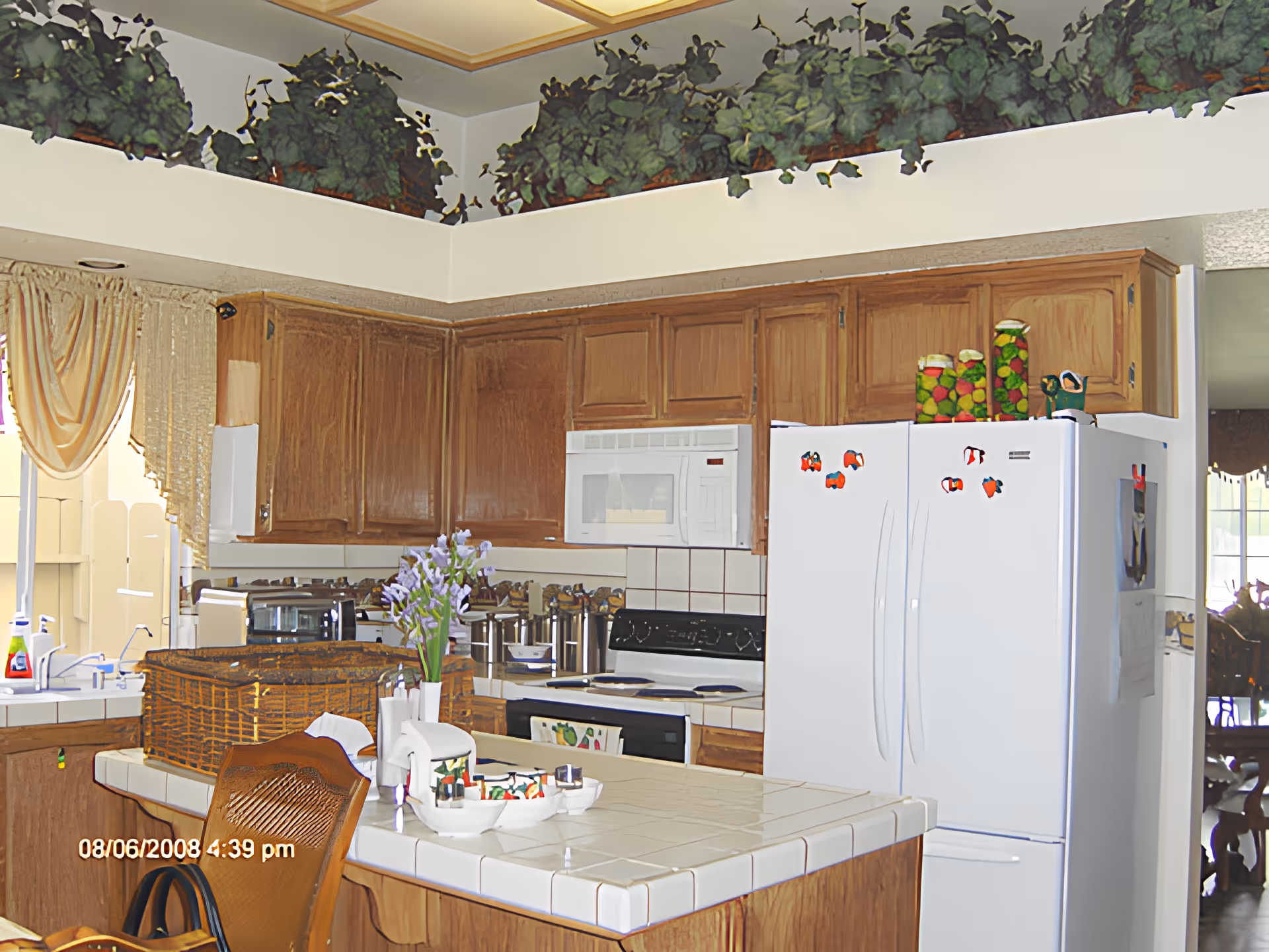 A kitchen with wooden cabinets, a white tiled countertop island with a vase of purple flowers, a white refrigerator with colorful magnets, a microwave above the stove, and decorative plants on a ledge near the ceiling.