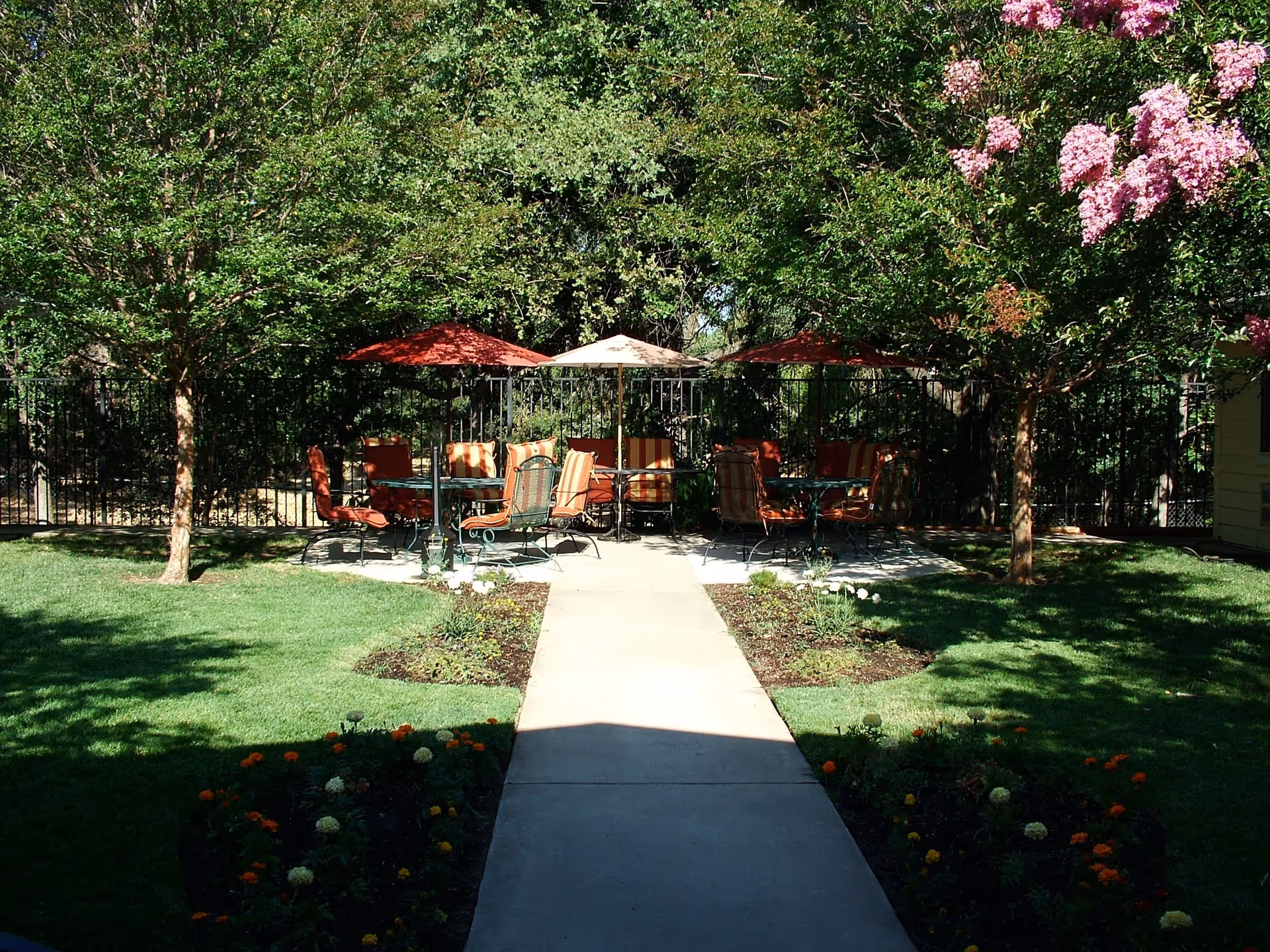 A peaceful outdoor patio area with several tables and chairs under red and beige umbrellas, surrounded by green trees and flowering plants. A concrete pathway leads to the seating area, with well-maintained grass and flower beds on either side.