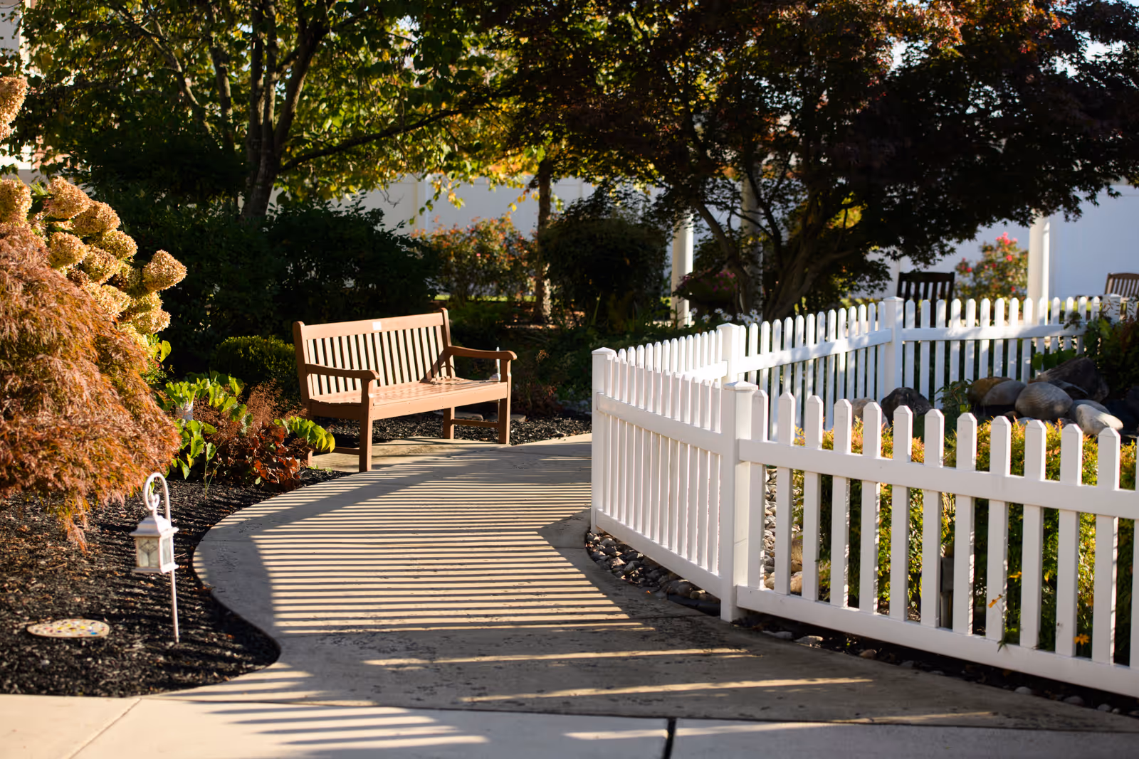 Sunlit garden path with a wooden bench beside a white picket fence and landscaped plants.