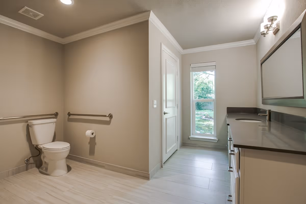 A spacious bathroom with beige walls and light-colored tile flooring. The room features a white toilet with metal grab bars on the walls beside it, a long vanity with a dark countertop and an undermount sink, a large rectangular mirror above the sink, a wall-mounted light fixture, a closed door, and a tall window showing greenery outside.