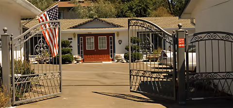 Open wrought iron gate leading to a driveway with a building in the background featuring a red double door entrance and an American flag displayed on the left side of the gate.