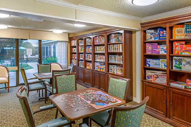 A cozy activity room with tables and chairs arranged for playing games and puzzles. One table has a partially completed jigsaw puzzle. Wooden shelves along the wall are filled with board games and puzzles. Large windows and glass doors provide a view of an outdoor garden area.