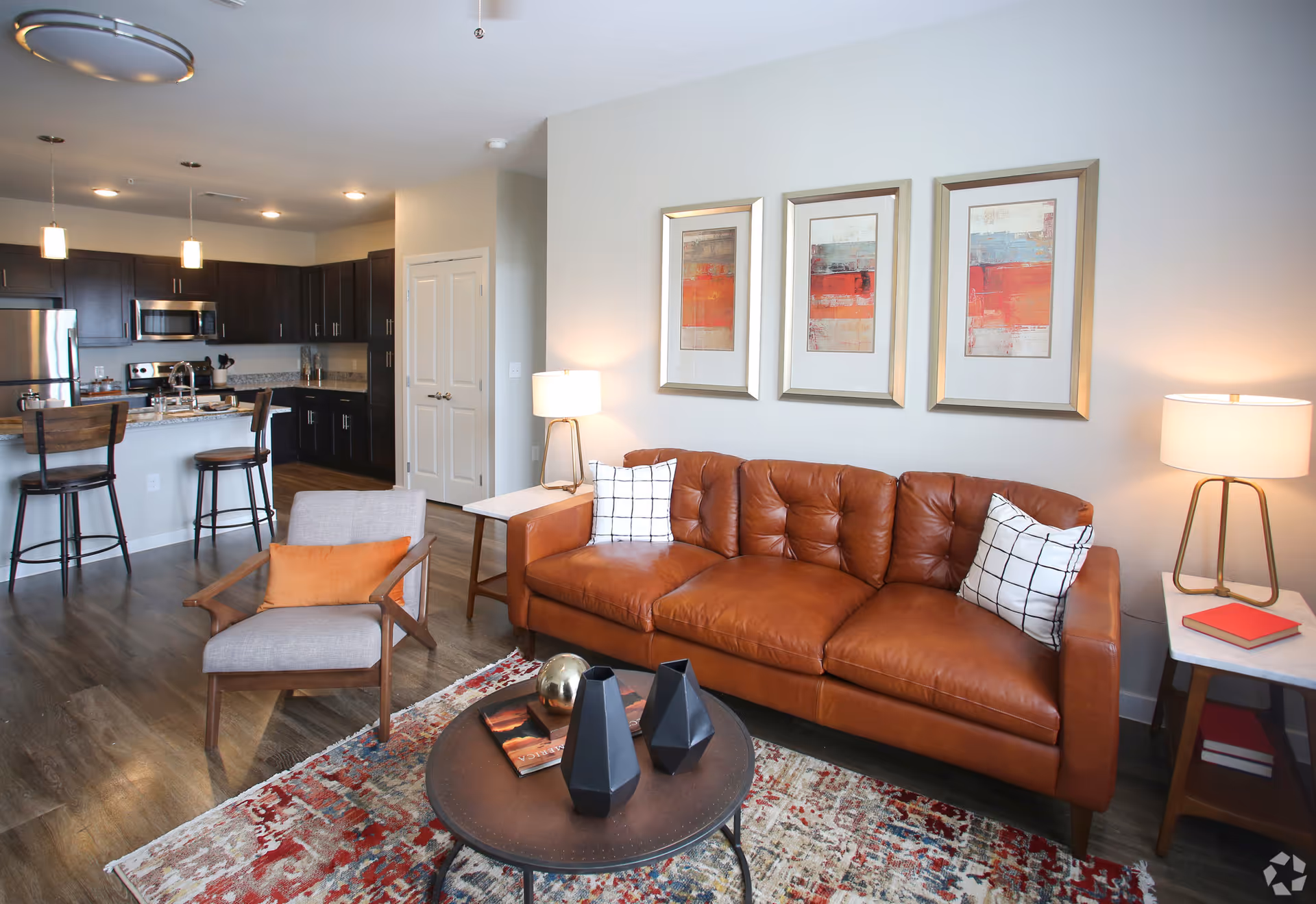 A modern living room with a brown leather sofa adorned with two white pillows featuring black grid patterns. Next to the sofa are two side tables with lamps and a red book on one of the tables. In front of the sofa is a round coffee table with decorative items and books on a colorful patterned rug. To the left, there is a gray armchair with an orange pillow and a kitchen area with dark wood cabinets, stainless steel appliances, and two bar stools at a counter.