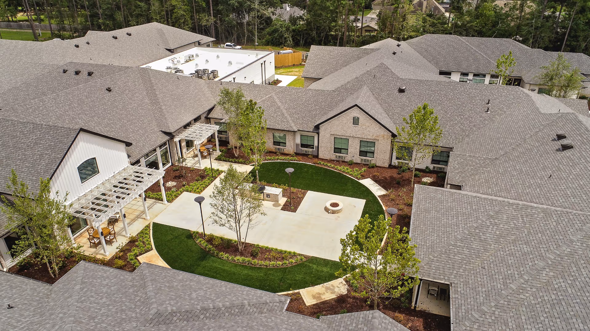 Aerial view of a senior living facility courtyard with a circular green lawn, concrete walkways, outdoor seating areas under pergolas, trees, and surrounding buildings with gray shingled roofs.