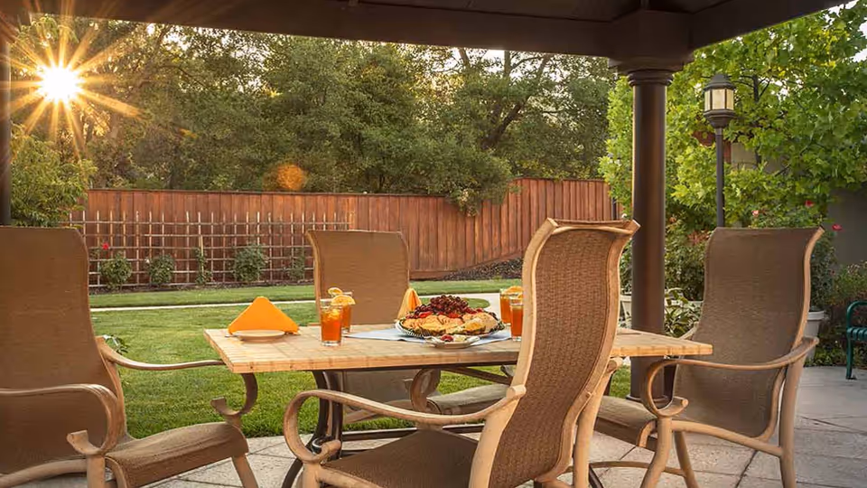 Outdoor patio area with a wooden table set for snacks and drinks, surrounded by four cushioned chairs. The sun is shining through trees in the background, with a wooden fence and green lawn visible.