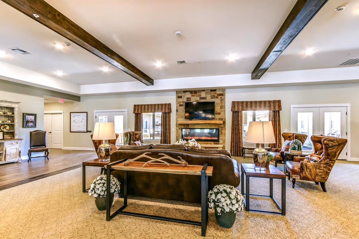 Spacious communal living room with sofas, armchairs, side tables and lamps facing a stone fireplace with a TV mounted above and exposed ceiling beams.