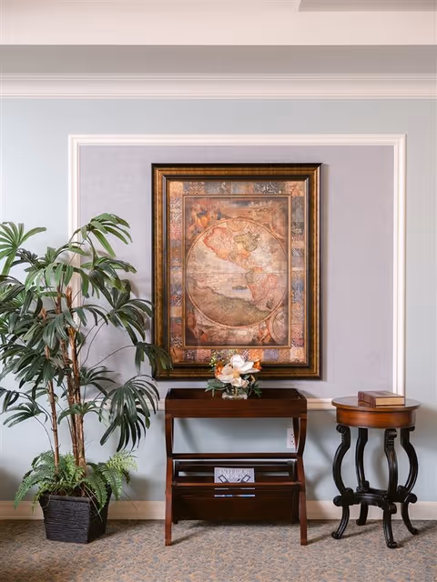 An interior corner of a room featuring a large framed vintage map on the wall above a dark wooden console table with a floral arrangement. To the left of the table is a tall potted plant with broad green leaves, and to the right is a small round wooden side table with a book on top. The wall is painted light blue with white trim, and the floor is carpeted.