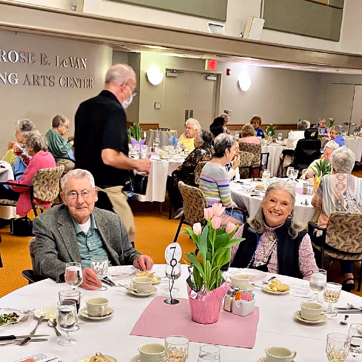 A dining room at Applewood Village with elderly residents seated at round tables covered with white tablecloths. The tables have cups, glasses, plates with food, and a centerpiece of pink tulips. A man wearing a mask and black shirt is serving coffee. The room is well-lit with wall sconces and has a sign on the wall that reads 'ROSE E. LeVAN ARTS CENTER'.