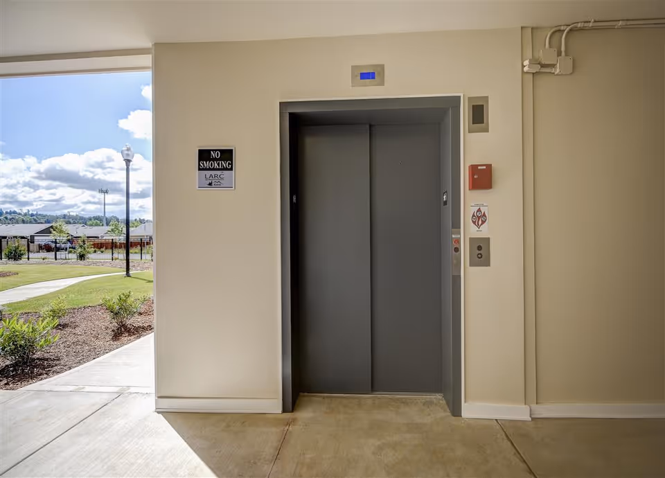 Closed elevator doors in a beige hallway with a 'No Smoking' sign on the left wall and an outdoor view with a pathway, grass, and lamp posts visible through an open area to the left.