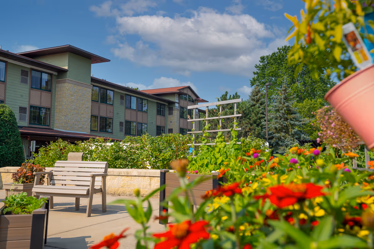 Outdoor garden area at Meadows on Fairview with colorful flowers in the foreground, a bench, planters, and a multi-story building in the background under a partly cloudy sky.