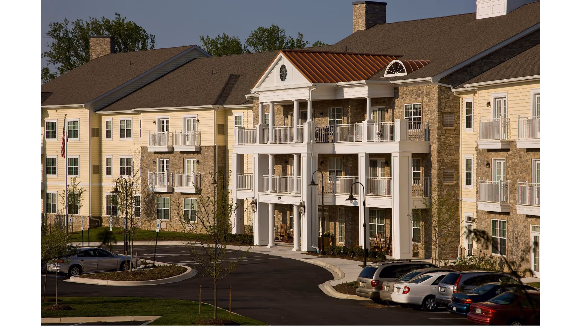 Exterior view of a senior living facility building with three stories, featuring a combination of stone and light yellow siding, balconies, and a covered entrance with white columns. Several cars are parked in front, and there are small trees and landscaped areas around the driveway.