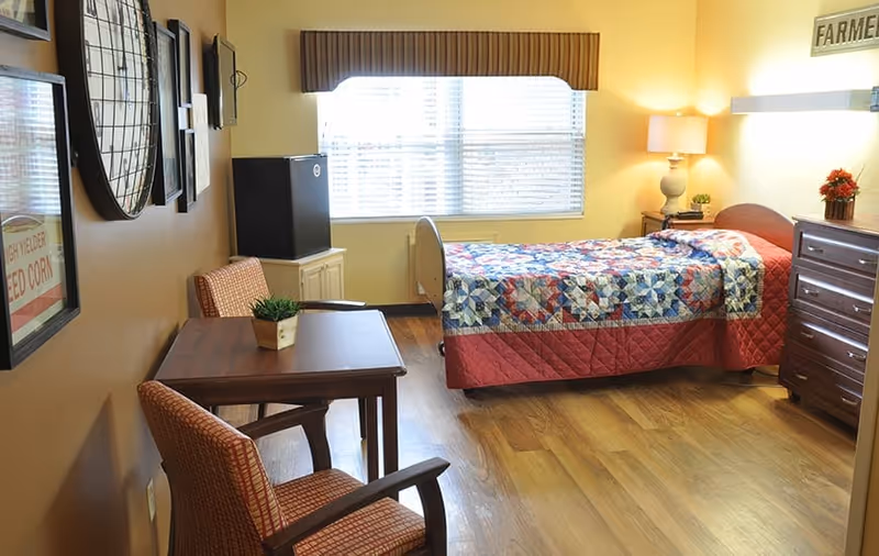 A cozy bedroom in a senior living facility with a single bed covered by a colorful quilt, a wooden dresser with a small plant and flowers, a bedside table with a lamp, a small table with two chairs, and a window with blinds letting in natural light.
