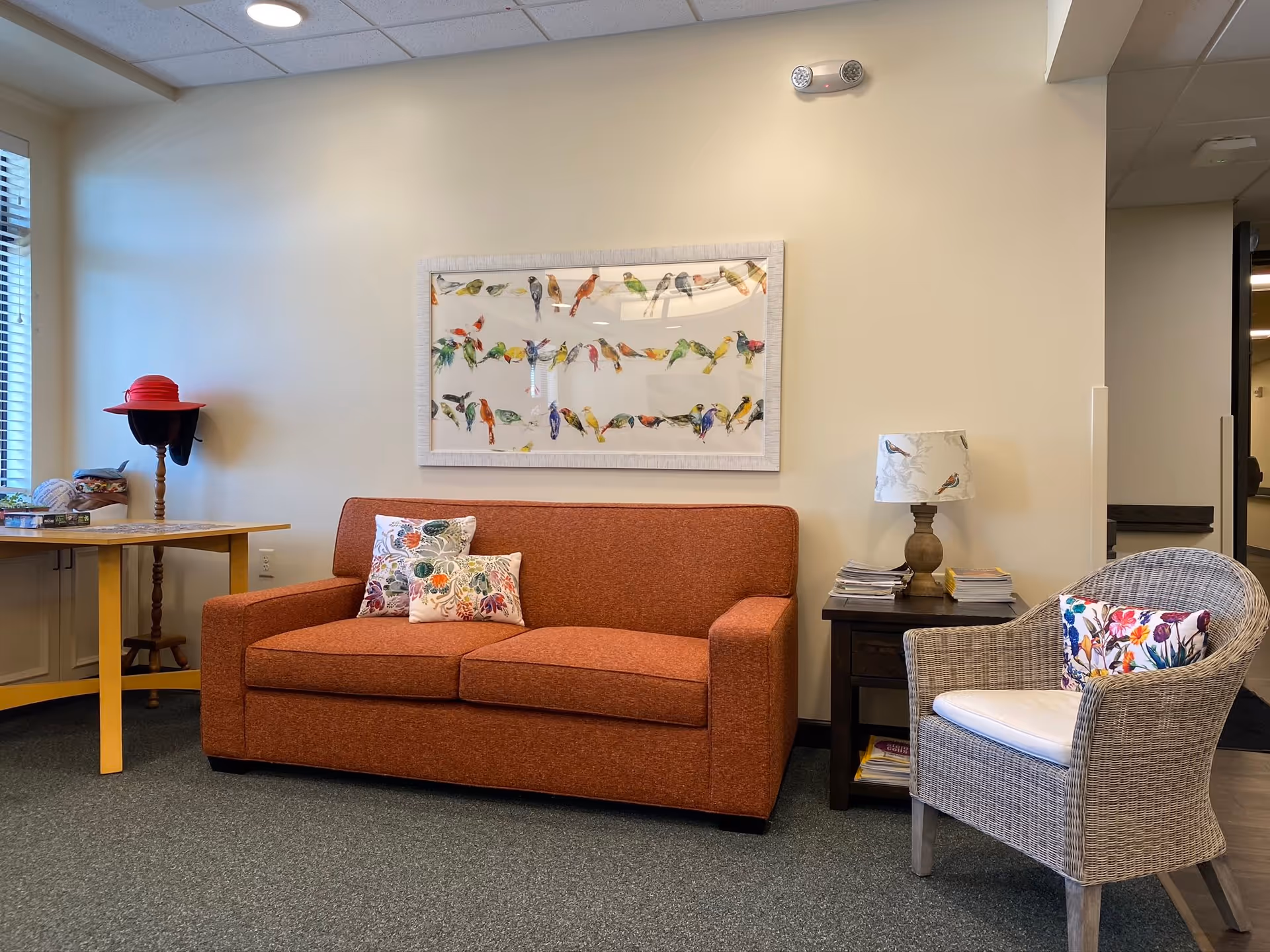 A cozy seating area in a senior living facility with an orange sofa adorned with floral pillows, a wicker chair with a floral cushion, a side table with a bird-patterned lamp and stacks of magazines, a wooden table with a red hat on a stand, and a framed artwork of colorful birds on the wall.